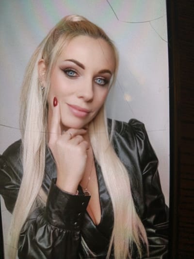 The image displays a head-and-shoulders portrait of a woman with long, straight, blonde hair, which appears to have a subtle pinkish tint towards the ends. Her hair is styled partly up in a high ponytail or half-ponytail, with some strands framing her face. She looks directly at the camera with a confident yet soft expression, featuring a slight smile.
Her eyes are a striking bright blue, accentuated by prominent dark winged eyeliner and what appears to be warm-toned eyeshadow. Her eyebrows are dark and well-defined. Her lips are full and have a natural pink or nude color. Her complexion appears smooth and well-made-up.
She is wearing a dark, possibly black, glossy or leather-like top or jacket with a V-neckline, revealing some cleavage. The garment features distinctive puffed sleeves, and buttons are visible on the cuff of her right sleeve. A delicate, thin necklace with a small, circular pendant hangs around her neck.
Her right hand is raised, with her index finger gently touching her right cheek just below her eye. Her fingernails are painted a dark red or maroon color. A simple band-style ring is visible on the ring finger of her right hand.
The background is light-colored and appears out of focus, suggesting a shallow depth of field. There are several faint lines and what look like cracks, particularly noticeable in the upper right quadrant of the image. These imperfections, along with a subtle screen-like texture, suggest that the image itself might be a photograph of a digital display or screen rather than a direct digital file. The lighting is soft and evenly illuminates her face. No text is visible in the image.