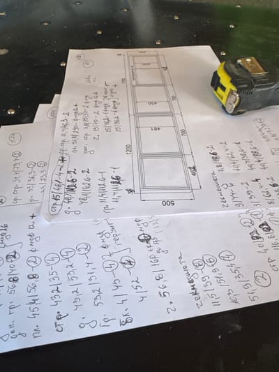 This image depicts a close-up, overhead view of several pieces of paper with handwritten notes and diagrams spread across a dark, textured surface, possibly a workbench or table. The papers appear to be technical drawings and calculations.
One sheet shows a schematic with dimensions, including measurements like 500, 450, 400, 700, and 462, indicating it might be a plan for a structure or object with multiple compartments. Another sheet contains a list of alphanumeric codes, dimensions, and symbols, such as "стр. 15/481/46.3-2", "пл. 51,8/290-ф", and various numbers with superscripts and parentheses. There are also circular notations with numbers inside, like "15", "13", "2", "1", "4", "8", and "14".
A yellow and black tape measure is visible on the right side of the image, resting on the papers and the dark surface.
The handwritten notes are in Cyrillic script and appear to be technical or architectural specifications. Some phrases are partially visible, such as "доп. гр.", "м. 45,4 | 56,8 | 40 + 2", "гр. 11,4 | 112,6-2", "ч. 432 | 35-4", "д. 43,2 | 53,2 - 2", "гр. 53,2 | 31,4 - 4", "Вк- 4 | 45- 4 | 45", "2. 56,8 | 160 + 4", "2. 15/70-2", "15/46,2-2", "15/96,2-1", and "тек. смет".
The overall scene suggests an active work or planning session related to construction or manufacturing. The specific location in the city of Belene, Bulgaria, cannot be determined from the image itself, as there are no visible landmarks or identifying signs related to that geographical area.