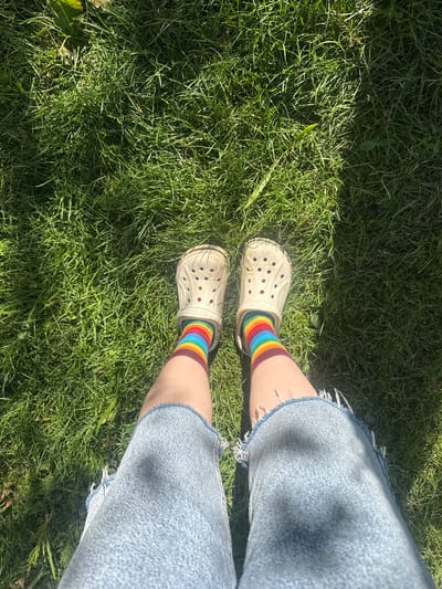 A pair of cream-colored Crocs are visible on a person's feet, standing on a patch of green grass. The Crocs are adorned with rainbow-striped socks, and the person is wearing distressed denim shorts. The grass is illuminated by bright sunlight, casting shadows. The scene appears to be outdoors, likely in a park or backyard setting. The location is identified as Kashka-Suu, Kyrgyzstan, though there are no specific visual cues in the image to confirm this location.