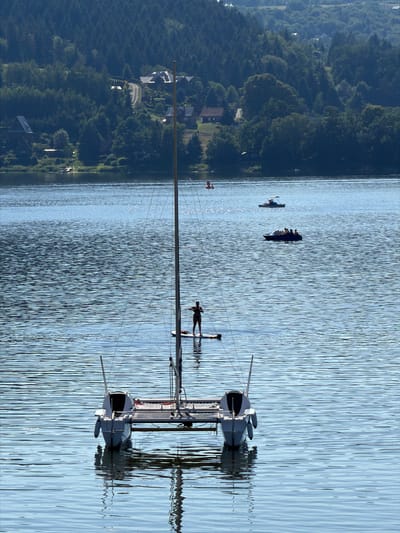 The image depicts a serene scene on a body of water, likely a lake, with a forested, hilly landscape on the left bank. Several boats are visible on the water, including a larger sailboat with a mast prominently featured in the foreground. A person is visible on a smaller boat or raft further out on the water, possibly engaged in water sports. In the distance, other small boats with multiple occupants are also present, suggesting a recreational outing. The water is calm with visible ripples, reflecting the sky and the surrounding scenery. The overall impression is one of a tranquil summer day. The forested hills dotted with houses suggest a rural or semi-rural location. The lighting indicates it is likely daytime, possibly late morning or afternoon, with clear weather. The reflection of the boats and landscape in the water adds to the picturesque quality of the scene. No visible text is present in the image.