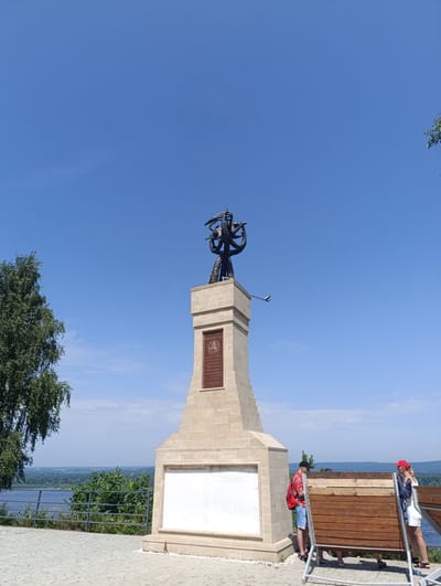 This is a full-shot, eye-level outdoor photograph taken on a bright, sunny day with a clear blue sky. The main subject is a tall, light-colored stone monument with a dark bronze statue on top. The statue depicts a figure with multiple arms holding various objects, appearing to be in a dynamic pose. Below the statue, on the monument, there is a dark plaque with an inscription and a relief image. Further down the monument is a large, white informational panel with text.
To the left of the monument is a large tree with green leaves. In the foreground, a paved area with light-colored interlocking stones is visible. A metal railing runs along the edge of this area, overlooking a body of water (likely a river) and a distant, tree-covered landscape.
On the right side of the frame, two people are interacting with a wooden structure. One person, a man wearing a red shirt, black cap, and blue shorts, is standing and looking at the wooden structure. Another person, a woman wearing a white top, white shorts, and a red cap, is also standing and looking at the wooden structure, holding what appears to be a phone. The wooden structure is a bench or viewing platform with horizontal wooden planks and a metal frame.
The scene is set in the city of Solnechnaya Polyana, Russia, known for its picturesque views and historical significance. The monument itself is likely a memorial or landmark. The presence of tourists suggests it is a public attraction.
