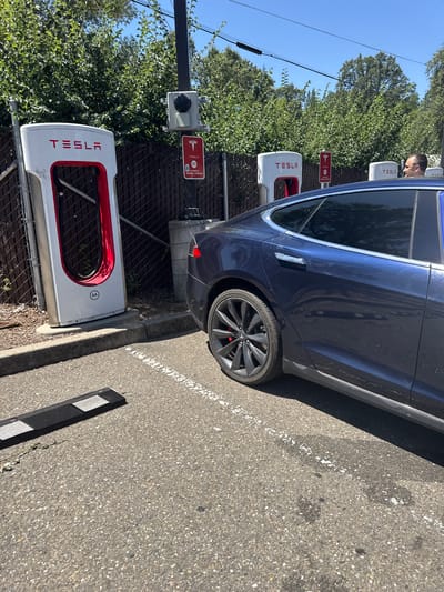 A dark blue Tesla Model S is parked at a Tesla Supercharger station in Placerville, California, United States of America. The car is positioned to the right of the frame, facing left. In the foreground, a black parking bumper with reflective strips is visible on the asphalt. Behind the car are three Tesla Supercharger units, two of which are clearly visible. The units are white with red accents and the Tesla logo. One charging cable hangs from the leftmost unit. A black fence runs behind the charging stations, separating them from a line of green trees and foliage. A man's head and shoulders are visible in the upper right corner of the frame, leaning out of the car. The sky is clear and blue. The scene is bathed in bright sunlight.