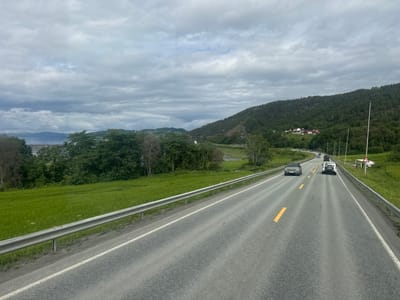The image depicts a rural highway scene on a cloudy day, likely in or around Verdalsøra, Norway. The perspective is from a vehicle moving along the road, looking forward. A two-lane asphalt road stretches into the distance, marked with yellow and white lines. A metal guardrail runs along the left side of the road, separating it from a vibrant green, grassy field. On the right side of the road, a similar guardrail is visible, with a steep, tree-covered hillside rising behind it.
Several vehicles are present on the road. In the foreground on the right lane, a dark grey SUV is visible. Ahead of it, in the left lane, is a white SUV. Further in the distance, more vehicles are seen, suggesting ongoing traffic.
The environment is characterized by lush green vegetation, including fields and dense forests that cover the hills. In the far distance, a body of water, possibly a fjord or lake, can be seen, with hills and mountains beyond it under a heavily overcast sky. A few scattered buildings, appearing to be houses or farmsteads, are nestled on the hillside in the middle ground, with red roofs on some of them, a characteristic of Norwegian architecture. A traffic warning sign, triangular with a red border and a symbol of a falling rock or debris, is situated on the right side of the road. The overall impression is one of a serene, natural landscape with signs of human infrastructure and activity.