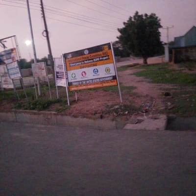 In Wuro Allahamdu, Nigeria, this image captures a street scene featuring a prominent sign for the International Rescue Committee (IRC) Emergency to Resilience (E2R) Project. The sign indicates support for Health, Nutrition, WASH (Water, Sanitation, and Hygiene), and Protection. The project is funded by the United States Government. In the background, a large tree and a building are visible, along with utility poles and overhead wires. The ground is covered with dirt and some scattered debris. The sky is overcast, suggesting twilight or a cloudy day. Other signs are also present in the background, though less legible.