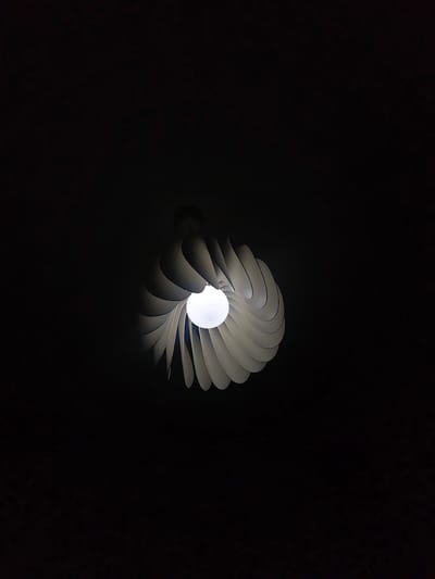 The image features a modern light fixture hanging from a dark ceiling. The fixture has a round lightbulb surrounded by white, petal-like structures arranged in a spiral. The lighting is dim, with most of the scene obscured by darkness. The exact location in Mosomishte, Bulgaria cannot be determined from the visual information provided.