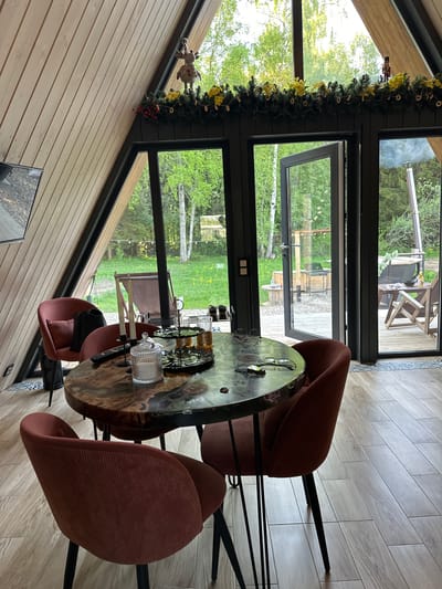 A residential interior in Малеевка, Russia, showcases a dining space. A round wooden table, adorned with glassware and serving pieces, is surrounded by four maroon-colored upholstered chairs with black metal legs. A wide window behind the table offers an outdoor view of trees and grass. The window frame is decorated with greenery and ornaments. Interior walls are lined with light-colored wooden paneling. Floor tiles are light grey. Through the open door a wood burning hot tub can be seen outside.
