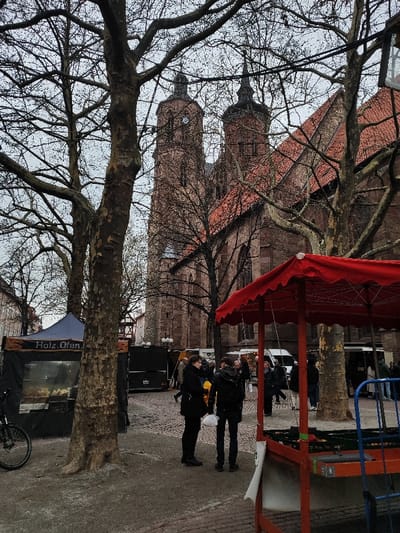 The image captures an outdoor market scene in Göttingen, Germany, with the St. Jacobi Church (Jacobikirche) dominating the background. The church is a large, reddish-brown brick structure with two prominent towers capped by dark spires and a red-tiled roof; the left tower features a visible clock face. The sky is uniformly overcast, and numerous large deciduous trees, bare of leaves, are scattered throughout the foreground and midground, their branches creating a dense canopy.

In the mid-ground, a market is in progress. On the left, a dark blue tent, possibly a food stall, displays a partial sign reading "Holz.Ofen." and features a transparent front panel revealing contents. A black bicycle is parked beside it. Towards the right, a large red market umbrella with scalloped edges covers a stall constructed with red framework and featuring blue metal bars on its right side. Several dark, mobile food trucks or market stalls and white vehicles are visible further in the background.

Multiple individuals are present, engaging in casual activity. Two figures stand prominently in the mid-ground: one individual in a dark coat holds a yellow item and a white bag, facing the other figure who wears a dark hooded jacket and dark pants. Other individuals are dispersed throughout the market area, either standing near stalls or walking, indicating a public gathering in a commercial setting.