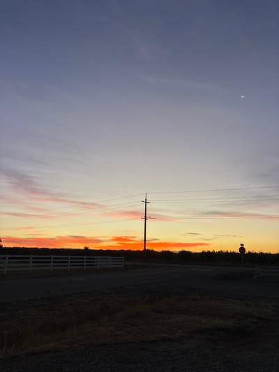 A serene twilight scene unfolds in Olivehurst, California, as a sliver of the moon hangs in the upper right quadrant of the sky, a pale crescent against the deepening blue. The sky transitions from a soft lavender near the top to hues of pale yellow and vibrant orange closer to the horizon, indicating the remnants of a recent sunset. Wisps of pink and orange clouds are scattered across the mid-sky, adding texture and depth.
Dominating the mid-ground, a telephone pole with multiple wires stretches across the frame, a stark silhouette against the colorful sky. A white fence runs horizontally along the left side of the image, bordering a dark, shadowy field. A road or paved area is visible in the lower half of the image, with a stop sign standing at a junction on the right side. The foreground is a mixture of dry grasses and gravel, suggesting a rural or semi-rural setting. The overall impression is one of quietude and natural beauty at the end of the day.