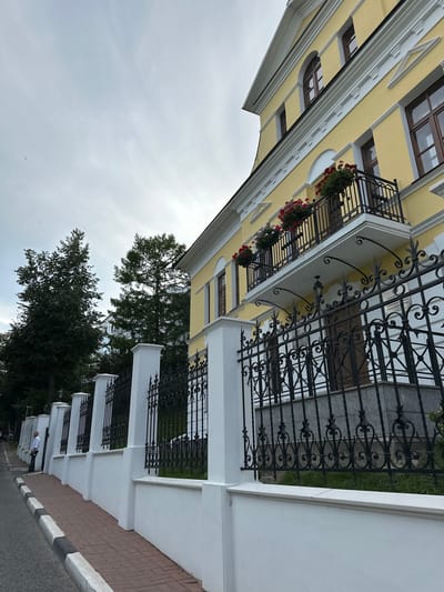 The image shows a yellow building with a white decorative cornice and arched windows. A black wrought-iron fence encloses the property, with white pillars supporting sections of the fence. There are red flowers in hanging baskets on a balcony. A person in a white shirt and dark pants stands on a brick sidewalk next to the fence. The sky is overcast. Trees are visible behind the fence and building.