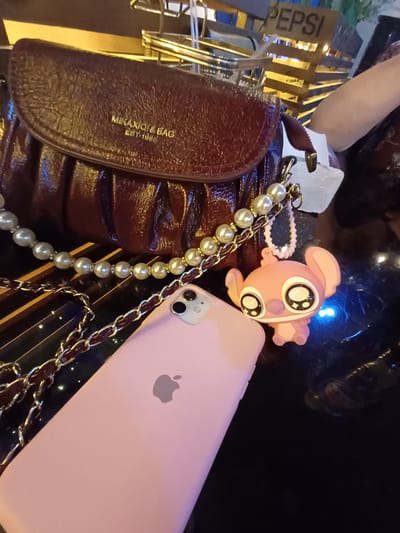 The image displays a collection of personal items arranged on a dark, reflective surface, possibly a table. Dominating the foreground are a dark brown, textured purse with gold lettering that reads "MIKAQIQI & BAG EST-1988" and a light pink iPhone. A pearl necklace is draped across the purse and a chain strap, which appears to be part of the purse or another accessory, is also visible. To the left of the purse, a partially obscured sign with the word "PEPSI" is visible, illuminated by yellow lights. A small, pink cartoon character charm with large eyes is attached to the pearl necklace. The overall scene suggests a personal moment, perhaps preparing to go out or showcasing items. The lighting is dim with some blue and yellow accents, hinting at an indoor setting at night. There are no clear indications of the weather or specific location beyond the "PEPSI" sign and the implied context of Nematjan, Uzbekistan. The image does not depict any overt emotions or a specific event, but rather a still life arrangement of everyday objects.