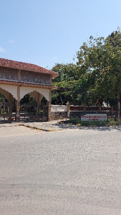 The image displays the entrance to Shree Swaminarayan Academy in Ziwa La Ng'ombe ward, Kenya. The perspective is from an asphalt road, textured and gray, which occupies the lower foreground. A prominent, ornate gate structure stands centrally, featuring two arched portals with closed black metal gates. The structure is built with light-colored, possibly stone or concrete, pillars and decorative elements, topped by a reddish-brown, patterned tiled roof. To the right of the main gate, a lower stone-block wall extends, also with reddish-brown decorative capping. In front of this wall, on a small patch of ground with sparse vegetation, a white rectangular sign with red text clearly reads "SHREE SWAMINARAYAN ACADEMY." Behind the gate and wall, a dense canopy of lush green trees and foliage is visible. The upper portion of the image is dominated by a clear, light blue sky, indicating bright, sunny weather conditions. No individuals, vehicles, or active events are present in the scene.