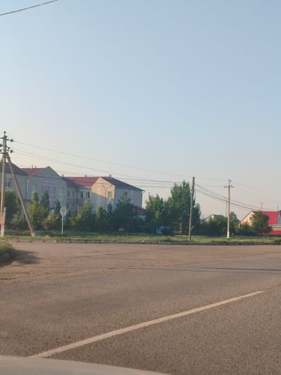 The image shows a street scene in the village of Postepnoy, Akzhayik District, West Kazakhstan Region, Kazakhstan. The foreground is dominated by a paved road with a white line marking. In the middle ground, there is a row of trees and vegetation along a curb. Behind the trees, a multi-story building with a red roof and light-colored facade is visible. To the right of this building, a smaller building with a bright red roof and a light pink facade is also present. Power lines and utility poles are scattered throughout the scene, indicating an urban or semi-urban environment. The sky is clear and blue, suggesting daylight. There are no people visible in the image. The overall impression is of a quiet, residential or administrative area.