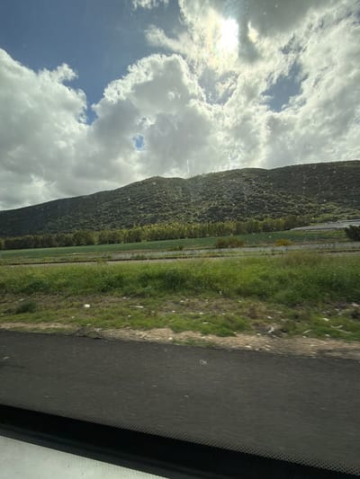 A photograph, seemingly taken from inside a vehicle, presents a landscape featuring a grassy verge, a forested hill, and a sky filled with clouds. The sky occupies a substantial portion of the image, showcasing a mix of blue sky and puffy white clouds, with the sun subtly shining through. The hill, densely covered with trees, stretches along the left side of the frame. The grassy verge, extending from the bottom of the image, appears unkempt with patches of brown and green. A road is visible in the foreground, with the edge of the vehicle's windshield and frame appearing on the right side of the image. The location is likely within Zvulun Regional Council, Israel, given the presence of characteristic terrain features. No people are visible in the shot.