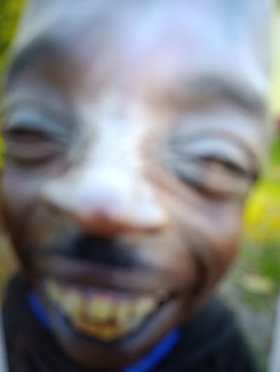 The image is a close-up, blurry, waist-up shot of a young Black male. He is outdoors, with blurry green foliage visible in the background. He is smiling broadly, showing his teeth, which appear discolored or possibly gold-capped. He is wearing a dark-colored garment with a blue collar visible. The image is highly out of focus, making it difficult to discern finer details of his features or the surrounding environment. There is no indication of the city of Krasivka, Ukraine, or any specific location.