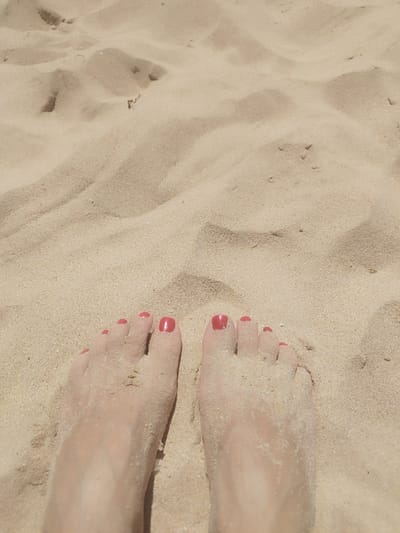 The image shows a close-up, overhead view of a pair of human feet partially submerged in sand. The feet are positioned in the foreground, with the toes pointing upwards. The skin of the feet is pale and dusted with fine sand. The toenails on both feet are painted a bright coral-red color. The background consists of undulating sand dunes, with patterns created by the wind. There are no other people or objects visible in the scene. The lighting suggests a bright, sunny day, typical of a beach environment. Given the context provided, this scene is likely located on one of the beaches in Rota, Spain, such as Playa de la Costilla or Playa de los Bateles, known for their sandy shores.