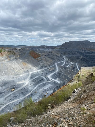 The image depicts a large open-pit mine located in Satka, Russia. The mine features terraced layers with winding roads traversing the levels. Several pieces of heavy machinery, possibly excavators or trucks, are visible on the roads. The pit walls are primarily gray in color with some areas of orange and brown discoloration. Overhanging the mine is a cliff covered in loose stones and some scrub vegetation. The sky is filled with gray clouds.
