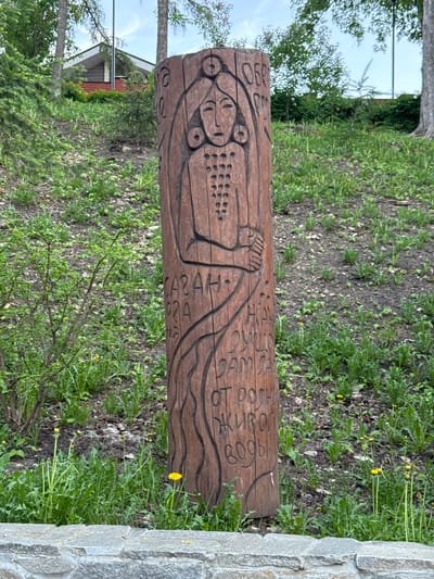 The image depicts a carved wooden log, situated in a grassy area, with a stone wall partially visible on the right side. The log features carved figures and script. The scene is located in Krasny Klyuch, Russia.