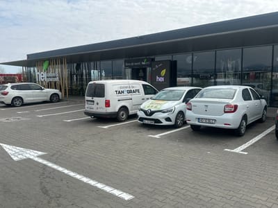 The image captures an outdoor scene at the "hei coffee & gourmet" establishment, located at Strada Mureșului 3, Orăștie, Hunedoara County, Romania, under an overcast sky.
The foreground consists of a grey interlocking paver parking lot, marked with white painted lines for individual parking spaces and a prominent white arrow indicating forward vehicular movement.
The background features a modern, single-story commercial building with a dark-colored roofline and extensive glass windows reflecting the surroundings. A section of the facade is decorated with vertical wooden slats. Prominent signage on the building includes the "hei coffee & gourmet" logo with a green leaf design and the text "hei coffee & gourmet," along with a sign above an entrance stating "Bine ați venit!" (Welcome!). The interior, visible through the glass, shows tables and chairs.
Four distinct vehicles are parked in marked spaces:
1. On the far left, a white BMW X3 SUV is parked, oriented towards the right. A partial license plate "P 473" is visible.
2. Adjacent to the BMW, a white Volkswagen Caddy panel van is parked, oriented with its rear facing the viewer. Its side panels display text, including "-atelierul de- TAHGRAFE -verificări tahografe digital / analog". A partial license plate "CJ 67" is visible.
3. Next to the van, a white Renault Clio hatchback is parked, oriented towards the viewer at a slight angle. The vehicle's hood and side feature green branding, including the word "DRUMAG". The license plate is "VL 22 BMH".
4. To the right of the Renault, a white Dacia Logan sedan is parked, oriented with its rear facing the viewer. Its license plate is "HD 12 BLC".
A portion of a fifth, darker vehicle is visible on the far right edge of the frame.
No individuals are present or visible within the captured scene. There are no ongoing actions or interactions.