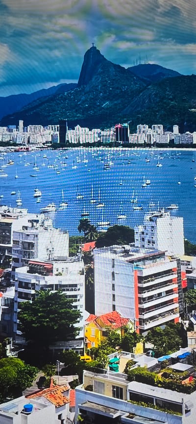 The image is a scenic aerial view of a cityscape with a prominent mountain in the background. The mountain has a statue of Jesus Christ at its summit, identifiable as Christ the Redeemer in Rio de Janeiro, Brazil. In the middle ground, a large body of water is filled with numerous sailboats and other vessels. The foreground depicts a dense urban area with various buildings, including modern high-rises with balconies and red-striped facades, as well as smaller houses with red-tiled roofs and lush green trees. The sky above is partly cloudy, with rays of light breaking through. The overall scene suggests a vibrant coastal city with a rich natural landscape.

The city of Lippstadt, Germany, is not depicted in this image.