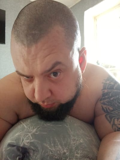 A bald man with a beard is lying on a pillow with a light-blue floral pattern. The man's left arm is visible, displaying a tattoo. The background is blurry, with a window visible on the right and a dark object, possibly a television, on the left. The man is looking down at the camera, with a furrowed brow. The location in Chulman, Russia is not discernible from the image.
