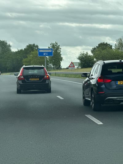 The image shows a perspective from inside a moving vehicle on a highway, looking towards the road ahead. A dark-colored Volvo station wagon, with a Dutch license plate "05-LVN-3", is in the left lane. To the right, in the right lane, is a dark blue BMW X1 SUV with a Dutch license plate "2-Z". Both vehicles are traveling away from the viewer.
In the background, a blue highway sign indicates "P+R Steenwijk" and the number "6". Further to the right, a triangular warning sign with a silhouette of a deer and the text "BBB LEVERT" and "2 KM" with an upward arrow is visible. The environment consists of a multi-lane asphalt highway bordered by green trees and vegetation under a cloudy sky. The location is on a highway in or near Tuk, Netherlands.
