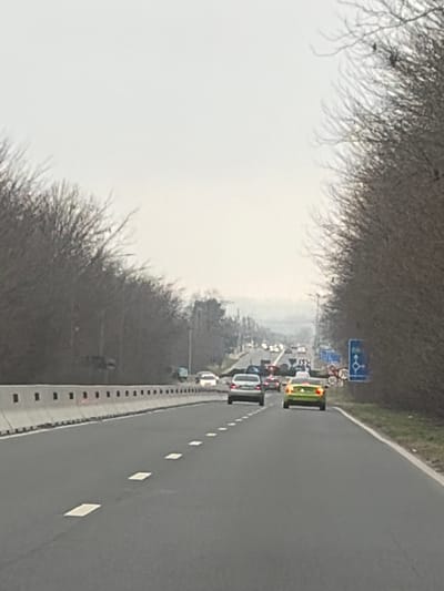 The image displays a multi-lane asphalt highway, likely a dual carriageway, in the vicinity of 23 August, Romania, observed from the perspective of a moving vehicle under an overcast sky. The road surface features white dashed lane markings. To the left, a concrete barrier with integrated black markers separates the roadway from a dense line of leafless deciduous trees. To the right, a narrow grassy shoulder precedes a similar dense line of leafless deciduous trees.

Traffic is visible ahead in the midground, with vehicles appearing to be slowing or queuing. In the immediate midground, a light grey sedan is positioned in the left lane, and a bright lime green sedan is in the right lane, both exhibiting illuminated red brake lights. Multiple other vehicles are discernible further down the road, some with illuminated brake lights.

On the right side of the road, a blue rectangular sign is affixed to a pole. This sign features a white circular arrow symbol indicating a roundabout. Below this symbol, white text is clearly visible, stating "23 AUGUST," with a circular speed limit sign indicating "70" partially visible underneath. The background reveals distant hills or elevated terrain, partially obscured by atmospheric haze. No individuals are clearly visible. The bare trees suggest a winter or early spring season.