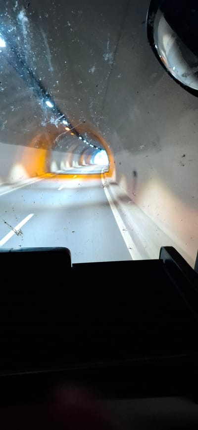 The image is a view from inside a vehicle traveling through a brightly lit tunnel. The tunnel walls are curved and concrete, with rows of lights visible along the ceiling, illuminating the roadway ahead. The road surface is smooth and gray, with white lane markings. The interior of the vehicle, including part of the dashboard and a side mirror, is visible in the foreground, along with a dirty windshield. The overall impression is of movement through a man-made structure. The location in the city of Blatino, Bulgaria is not discernible from the image.