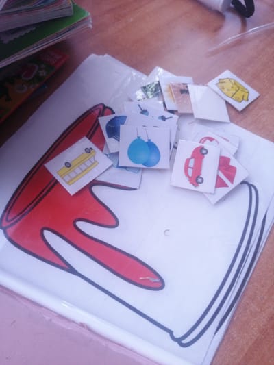 The image shows a close-up view of a table covered with various items, likely for a children's activity. A stack of colorful books or folders is visible in the upper left corner. In the center, a large white sheet of paper with a drawing of a red shape outlined in black is partially covered by a scattering of small, square cards. These cards feature simple cartoon-like illustrations of objects, including a yellow school bus, blue balloons, a red car, and a yellow jacket. Some of the cards are slightly bent or overlapping. The background is a wooden table surface. The lighting suggests an indoor setting, possibly during daylight. The overall impression is one of a playful and educational activity, perhaps for young children. No people are visible in the image. The scene appears to be set in Dubrovka, Russia, as indicated by the provided location context, though there are no explicit visual cues in the image to confirm this. There is some faint text visible on the spine of one of the books in the upper left corner, but it is too blurry to read.