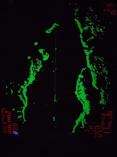 The image displays a green-on-black radar screen, likely from a marine vessel or weather monitoring station. Bright green, irregular, and fragmented shapes dominate the black background, representing radar returns which could indicate landmasses, islands, or significant weather formations. A prominent, thin vertical green line dissects the screen, possibly a bearing line or part of the radar's active sweep.
Various text elements in red and white provide operational information and settings. On the left, "DISP STD BRILL1" heads a list including "EBL," "VRM," "NAV," "HL," "RNGS," and "DATA," each with red indicator bars suggesting they are active or set. Below these are "PANEL BRILL" and "ECHO COLOR," with "BACK COR" highlighted in blue.
On the right, "DISP STD ECHO1" is visible, along with specific settings such as "FTC OFF," "IR 1," "ES 410," and "NR OPP." Further options like "REL 2SCAN EAV" and "TRAILS OFF CONTRAST 1" are shown, with "TRAILS OFF" indicating that historical radar data is not being displayed. "AUTO TUNE," "GAIN," "A/C SEA," and "A/C RAIN" are also listed, likely related to radar sensitivity and clutter control. "LOCAL" appears in the top right corner.
The scene captures an active radar system providing real-time data, typically used for navigation, collision avoidance, or weather assessment. The fragmented green returns suggest a detailed and possibly complex environment being scanned. No specific time of day or weather is directly inferable from the display, which is characteristic of instrumental readings. The location context of Kudryashovo, Russia, is not visually discernable from the abstract radar map.