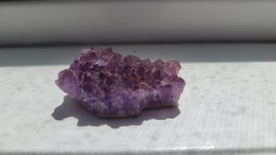 A close-up, side-view, low-angle shot shows a cluster of purple amethyst crystals on a white textured surface. The crystals are rough, jagged, and have a faceted appearance. They are illuminated by natural sunlight, casting a distinct shadow to the left of the cluster. The background is out of focus, showing a muted grey horizontal band above a lighter grey area, possibly indicating a windowsill or a shelf against a wall. The image is taken indoors. There is no indication of location within Terrebonne, Canada.