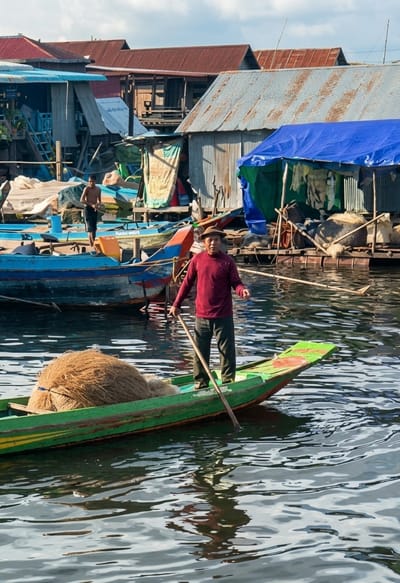 A man wearing a straw hat and a maroon long-sleeved shirt stands in a green and yellow boat, holding a wooden oar. The boat contains a large pile of coiled rope or net. In the background, a village is built on the water, with houses constructed on stilts. The buildings have corrugated metal roofs and wooden walls, and some are draped with colorful tarps. Other boats are moored in the water. Another young man, bare-chested, stands in a blue boat to the left. The scene depicts a waterside community, likely the floating village of Kampong Phluk in Soutr Nikom, Cambodia, with people engaged in daily activities on the water.