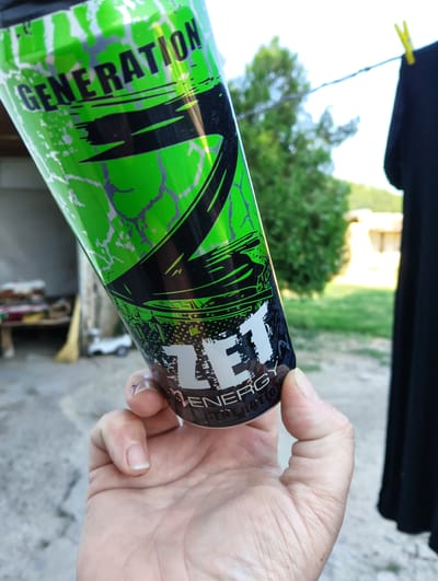 A close-up, slightly tilted shot shows a person's hand holding a tall, green and black energy drink can. The can has a jagged, abstract design with streaks of white. Large, bold, black text across the top reads "GENERATION". Below this, a white logo with black outlines contains the letters "T", "L", and "F", with "ENERGY GENERATION" written in smaller text underneath. The background is softly blurred, revealing an outdoor setting. To the left, a light-colored wall and a wooden shelf with items on it are visible. Further back, a green tree stands against a bright sky. To the right, a black garment hangs on a clothesline, secured by yellow clothespins. The lighting suggests it is daytime, possibly late afternoon, with bright sunlight. The overall scene appears to be a casual, outdoor environment, perhaps a backyard or patio area. The focus is on the energy drink can, suggesting the person is about to consume or is holding it for display. The image is sharp on the can and the hand, with the background out of focus. The provided location context of "Баяндай, Kazakhstan" indicates the geographical setting.