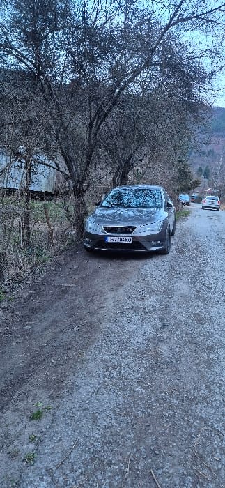 A grey SEAT Leon X-PERIENCE, bearing Bulgarian license plate CA 7204 KO, is stationary on the left side of an unpaved gravel and dirt road in Tompsan, Bulgaria. The vehicle's windshield displays a layer of frost or condensation. To its immediate left stands a large, leafless deciduous tree, partially obscuring a dilapidated wooden structure. The road surface is composed of gravel and compacted dirt, with sporadic patches of green vegetation along the verge. In the distant background, the road continues, revealing two additional vehicles: a dark sedan or hatchback followed by a white SUV or station wagon. Forested hills or mountains are visible under an overcast, light blue-grey sky. The scene is illuminated by diffused natural light, consistent with twilight or early morning conditions. No individuals are present.