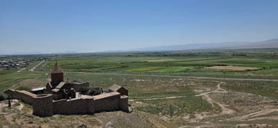 The image captures a panoramic view of the Khor Virap monastery complex, situated in Lusarat, Armenia, on a clear, sunny day. The monastery, constructed primarily of stone, features a prominent conical roof atop its central church. Stone walls surround the complex, with a few smaller structures and a cross visible within the walled area. Several individuals are present within the monastery grounds, seemingly exploring or visiting.

Beyond the monastery, the landscape unfolds into a vast expanse of agricultural fields, displaying a patchwork of green and yellow hues, indicative of various crops. A road or pathway winds through the fields, and a fence runs parallel to it in the middle ground. In the distance, a town or village with scattered buildings is visible to the left of the monastery, with a road leading towards it. Further back, rolling hills and mountains form the horizon under a clear blue sky. The terrain leading up to the monastery is dry and rocky, with sparse vegetation. The overall scene is one of historical significance set against a backdrop of rural agricultural life and natural mountainous scenery.