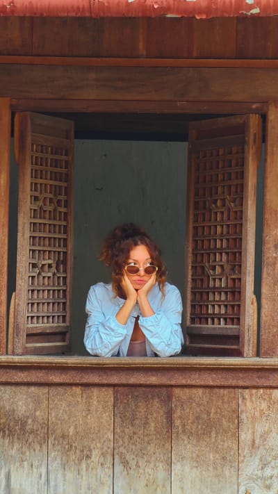 A medium shot depicts a woman seated in a traditional wooden window in Sà Phìn Commune, Vietnam. The woman has curly red hair, wears round sunglasses, and a light blue shirt. She rests her chin in her hands, gazing out the window. The window frame is dark wood, and the shutters are partly open, revealing a gray wall behind the woman. The window frame is textured, and the shutters have a square lattice pattern. The bottom part of the window has a panelled wood surface.