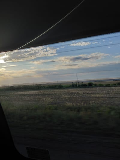 The image shows a view from inside a vehicle, looking out through a window at a rural landscape under a cloudy sky. In the foreground, the dark interior of the vehicle, likely the car's headliner or a sun visor, partially obscures the view. A thin, bright line, possibly a reflection or light streak, crosses the dark area diagonally.
The middle ground of the image features a broad, flat field, appearing to be agricultural land, possibly harvested. The texture of the field is a mix of dry grasses and stubble. A line of green vegetation, consisting of trees and shrubs, runs horizontally across the middle distance, separating the foreground field from the background. Beyond this vegetation, there are distant, hazy landscapes that suggest a very flat terrain, with what might be a body of water or a low-lying plain in the far distance. Utility poles are visible scattered across this distant landscape, indicating human presence.
The sky is filled with a variety of clouds, ranging from wispy cirrus to more substantial cumulus formations. The sun is visible on the left side of the frame, partially obscured by clouds, casting a soft, diffused light that illuminates the sky and the landscape. The overall impression is of a serene, expansive view of a rural area, likely captured during daylight, possibly late afternoon or early morning due to the angle of the sun and the quality of light. The image appears to have been taken while in motion, given the slight blur in the field and the streaks of light. The location mentioned, Молодёжный, Kazakhstan, is consistent with the observed landscape of flat, open fields and a wide sky.
