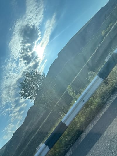 The image shows a road in Arzni, Armenia. A guardrail runs along the right side of the road, with a steep, grassy hillside rising to the left. The sky is blue with scattered clouds and the sun is shining brightly, casting rays of light across the scene. A tree with dense foliage is visible on the hillside. The road appears to be paved. The image is taken from a slightly tilted perspective.
