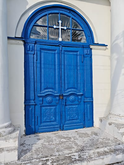 A blue arched double door with a cross in the transom stands at the entrance to a building in Мамонтово, Russia. The door has decorative carvings and is secured with a padlock. The building is white, with fluted columns on either side of the entrance. The ground is a worn, tiled surface. The scene is sunlit.