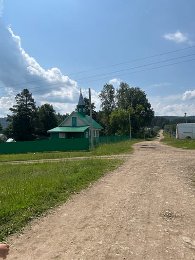 The image depicts a rural scene on a sunny day, likely in the village of Novomullakayevo, Russia. In the foreground, a dirt road winds into the distance, bordered by grassy verges. To the left, a vibrant green wooden fence encloses a property with a distinct green house featuring a steeply pitched roof and a pointed spire. Tall, mature trees surround the house and property. Further down the road, more trees line the path, and a white van is visible on the right side of the road. The sky is a bright blue with scattered, fluffy white clouds. A person's hand is partially visible in the lower left corner of the frame, suggesting they are holding the camera.