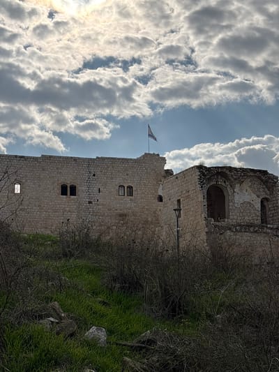 This image presents a forensic view of Migdal Afek (Antipatris Fortress) located in Rosh HaAyin, Israel. The primary subject is a multi-sectioned stone fortress constructed from light-colored, possibly limestone, blocks. The left section of the fortress appears largely intact, featuring a flat roofline and several rectangular windows. One window on the far left shows light through its opening, while others appear dark. A blue and white Israeli flag, bearing a Star of David, is flown from a flagpole positioned on the highest point of the fortress, near the center-right of the main structure. To the right of the main section, a more ruinous part of the fortress is visible, characterized by crumbling walls and large arched openings. The foreground consists of a sloping terrain covered in sparse, dry, dark-toned brush and patches of vibrant green grass, particularly prominent in the lower left. Scattered rocks and natural debris are present on the ground. A dark-colored, vertically oriented lamppost with a light fixture is situated in the mid-ground, positioned to the right of the main fortress section and in front of the ruined portion. The sky above is partially overcast, with a bright, diffused sunlight source visible through clouds in the upper left, and segments of clear blue sky towards the upper right. No individuals, explicit actions, or interactions are depicted.