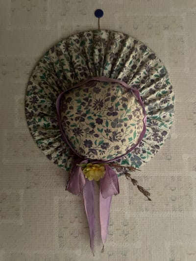 This media file features a small, decorative bonnet made of floral fabric. The bonnet is light-colored with a pattern of purple and green flowers. It has a ruffled brim and a gathered crown. A delicate purple ribbon is tied around the base of the crown, forming a bow. Attached to the bow are a small yellow fabric flower and a sprig of dried lavender. The bonnet is placed on a textured, light-colored surface that appears to be fabric or paper. A small, dark blue pin is visible on the left side of the image, slightly to the left of the bonnet's brim. The lighting suggests an indoor setting, possibly a craft room or a display table. The overall impression is one of delicate craftsmanship and a touch of rustic charm, with the lavender evoking a sense of Provence. The image is a still life, with no people or specific activity taking place beyond the arrangement of the object.
