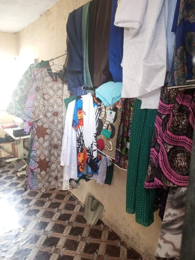 Garments hang on rods attached to a worn wall above a patterned tile floor. A sewing machine table sits in the background, left of frame. The clothing includes an assortment of colors and fabrics such as green, blue, brown, white, and floral patterns. The clothing hangs from a metal rod. There is also a tan bag on the floor by the wall.