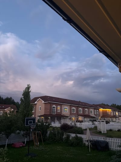 The image depicts a suburban residential area with several townhouses under a cloudy sky, likely at dusk. The townhouses are brick-faced with dark red roofs and have illuminated windows, suggesting occupancy. A white picket fence runs along the front of the properties, bordering a well-maintained lawn area. In the foreground, a basketball hoop stands on a black pole, with a red inflatable toy and a children's slide visible in the background. There are trees and bushes scattered throughout the scene, and a path is visible behind the fence. The overall impression is one of a quiet, family-oriented neighborhood in Baranovo, Russia.
