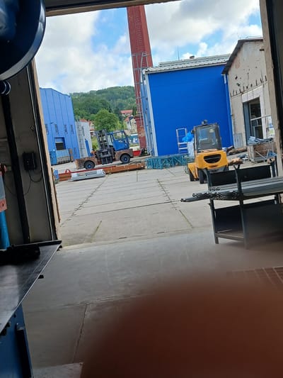 The image depicts an outdoor industrial area, likely a factory or construction site, viewed from within a building's open doorway. In the foreground, a metal table or workbench is visible on the left. The central area of the image shows a concrete yard with various industrial elements. A blue building with white-trimmed windows stands prominently in the middle ground. To its right, a small yellow forklift is parked. Further to the right, a more weathered, light-colored building with damaged facade is visible. In the background, a red brick chimney rises into a cloudy blue sky, and a lush green hill with scattered houses can be seen. The overall scene suggests a working environment with active construction or material handling operations. The context of Benešov nad Ploučnicí, Czechia, implies an industrial or commercial setting within that specific geographical location.
