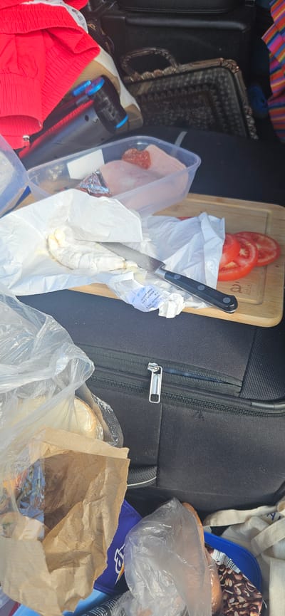 The image depicts a close-up, overhead view of the trunk of a vehicle, suggesting an outdoor setting potentially in Beňadikovce, Slovakia. In the foreground, a black suitcase with a silver zipper is visible. Next to it, a brown paper bag and a clear plastic bag contain food items, including what appears to be a loaf of bread. A blue plastic container and a white cloth bag are also present. In the middle ground, a wooden cutting board holds three sliced tomatoes and a piece of cheese wrapped in white paper, with a knife resting on it. A clear plastic container filled with sliced ham and salami is behind the cheese. In the background, a red garment, a dark-colored bag with a patterned design and a handle, and a striped fabric are visible. The overall scene suggests a prepared meal or picnic.