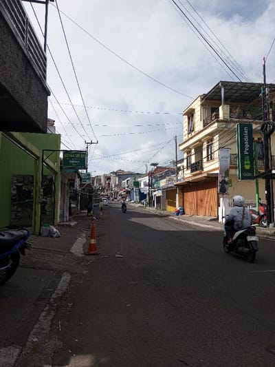 An urban street scene features a narrow road flanked by multi-story buildings on both sides. The buildings exhibit various architectural styles and storefronts, some with closed metal shutters. Overhead power lines crisscross the sky. A motorcycle with a single rider is moving down the street. An orange traffic cone is on the side of the road near a parked motorbike. The sky is partly cloudy, casting shadows on the street.