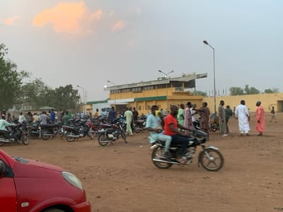 The image shows a scene outside a building in Sokoto, Nigeria, located at coordinates 13.049 latitude and 5.238 longitude. There are numerous motorcycles parked in an open dirt lot. Some people are gathered around the motorcycles, while others are walking away from the scene. Several individuals are riding motorcycles. There is a red car parked in the foreground. A building with a green and yellow facade is located in the background. There is a green and white sign above the entrance to the building. There are trees and shrubs scattered throughout the scene. The sky is overcast with a faint sunset glow. The environment is dusty, with an open dirt lot and some patches of grass. The overall atmosphere is casual and busy, with a mix of people and vehicles. The focus of the image is the gathering of people around the motorcycles, suggesting an informal event or a gathering. The building in the background suggests a public place or a sports facility. The image captures a moment of everyday life in Sokoto, Nigeria.
