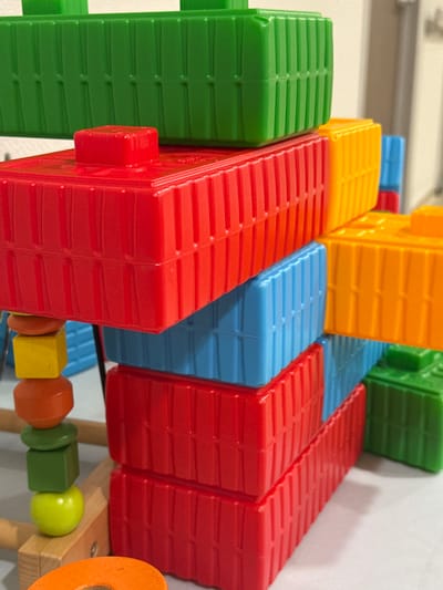The image shows a close-up of colorful plastic building blocks stacked in a complex arrangement. The blocks are primarily red, green, and blue, with some yellow and orange elements visible. The blocks appear to be toy construction pieces with a ridged texture. In the foreground, on the left side, there's a wooden toy with several colorful beads stacked on it, including a yellow cube and orange rounded shapes, with a green sphere at the bottom. The background is out of focus and appears to be an indoor setting with a light-colored surface. There are no people or animals in the image. The composition is a still life of toys. There is no discernible information in the image that would allow for a location in Sultanbeyli, Turkey.