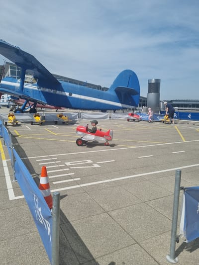 The image shows an outdoor scene at Stuttgart Airport in Echterdingen, Germany. A large blue and white biplane dominates the left side of the frame. In the center, a child wearing a green hat and jacket sits in a small red airplane toy. Several other airplane toys, primarily yellow, are parked nearby. An adult in a dark top is standing next to the biplane on the left. The ground is marked with yellow and white lines, suggesting a parking area or runway. A blue flag on a pole is visible in the foreground, and an orange traffic cone stands near it. In the background, airport buildings are visible. The sky is blue with scattered clouds.