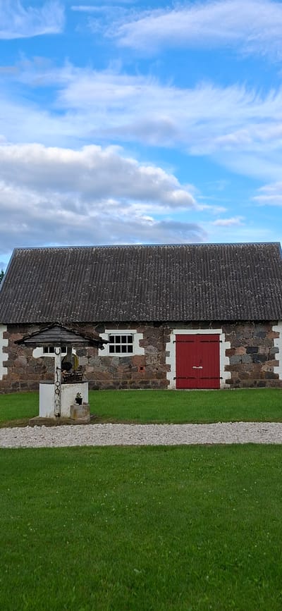 This is a photograph of a rustic stone building with a corrugated roof and red double doors, located in the village of Viru-Jaagupi alevik, Estonia. In front of the building is an old stone well with a wooden roof. The well is situated on a gravel path that leads to the building, and the area is surrounded by green grass. The sky above is blue with scattered white clouds. The building's facade is constructed from irregularly shaped stones of various colors, with white stone accents around the windows and doors. The windows are divided into multiple panes. The red doors have visible hinges and a latch. The well structure is white, with visible ropes and a mechanism for drawing water. A small potted plant sits on the well's base. The overall scene suggests a historical or rural setting.